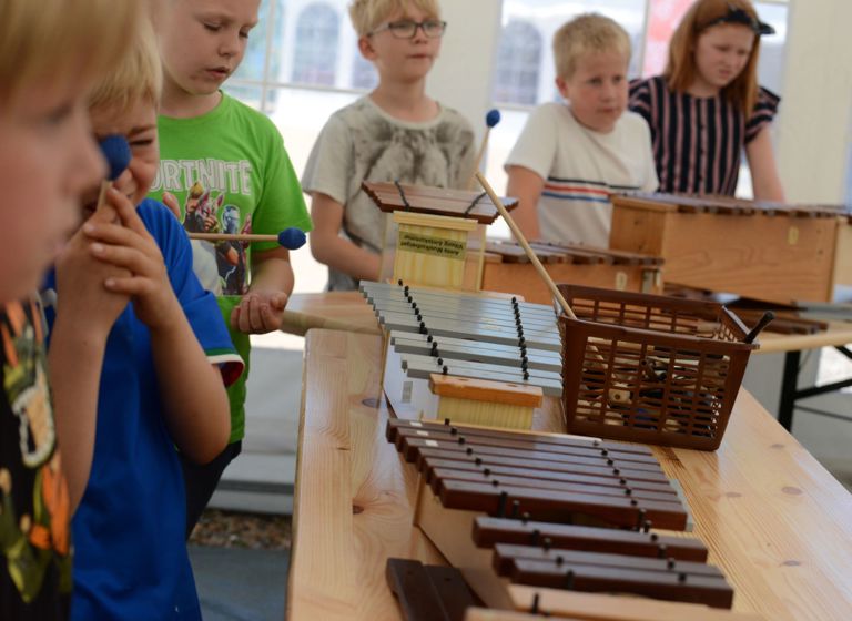 Børn står omkring et bord med forskellige slagtøjsinstrumenter, herunder xylofoner og andre mallet-instrumenter.