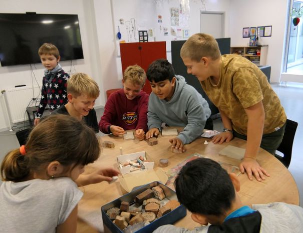 En gruppe børn er samlet omkring et bord og leger med ler. Børnene ser ud til at være fokuseret på opgaven, og nogle rækker ud efter ting i en kasse på bordet.