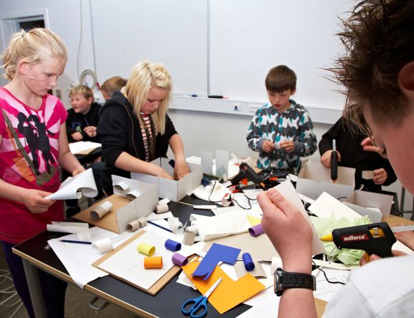 En gruppe børn engageret i en håndværksaktivitet ved et bord. Bordet er fyldt med forskellige håndværksmaterialer, herunder papir, sakse, limstifter og andre forsyninger. Børnene er fokuseret på deres opgaver, hvor de klipper og samler forskellige stykker papir og materialer.