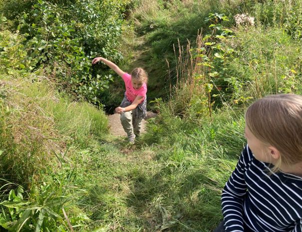 Et barn i en lyserød trøje og beige bukser går på en græssti omgivet af grønt. Et andet barn i en sort-hvid stribet trøje sidder til højre i billedet.