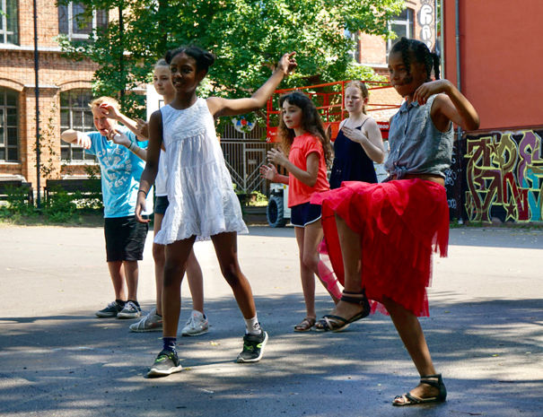 En gruppe børn leger eller danser udendørs i et solrigt område. Baggrunden inkluderer en murstensbygning, træer og farverig graffiti på en væg.
