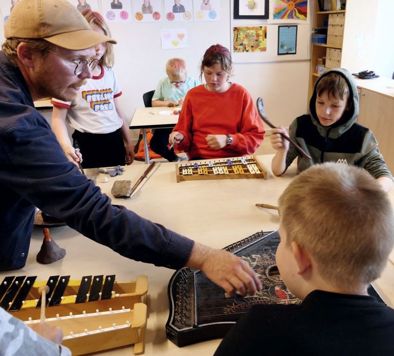 En gruppe mennesker i et klasseværelse spiller forskellige musikinstrumenter, herunder xylofoner og andre slagtøjsinstrumenter. En person står op og hjælper eller instruerer de andre.