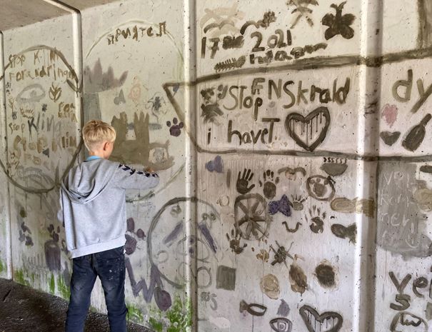 Et barn står foran en væg dækket med mudder-graffiti og tilføjer til graffitien.