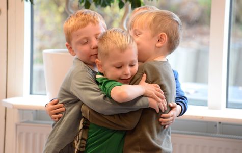 Fire børn står i et gruppekram. De er indendørs, med et vindue og noget grønt synligt i baggrunden.