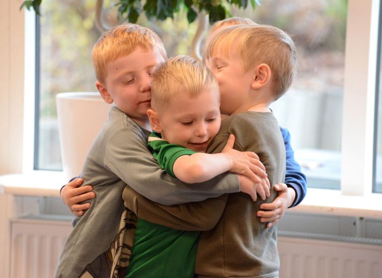 Fire børn står i et gruppekram. De er indendørs, med et vindue og noget grønt synligt i baggrunden.