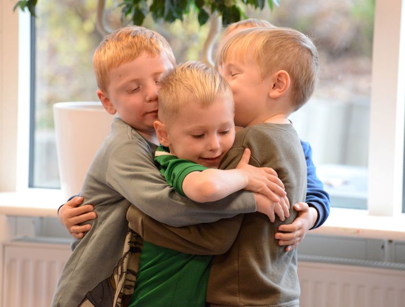 Fire børn står i et gruppekram. De er indendørs, med et vindue og noget grønt synligt i baggrunden.
