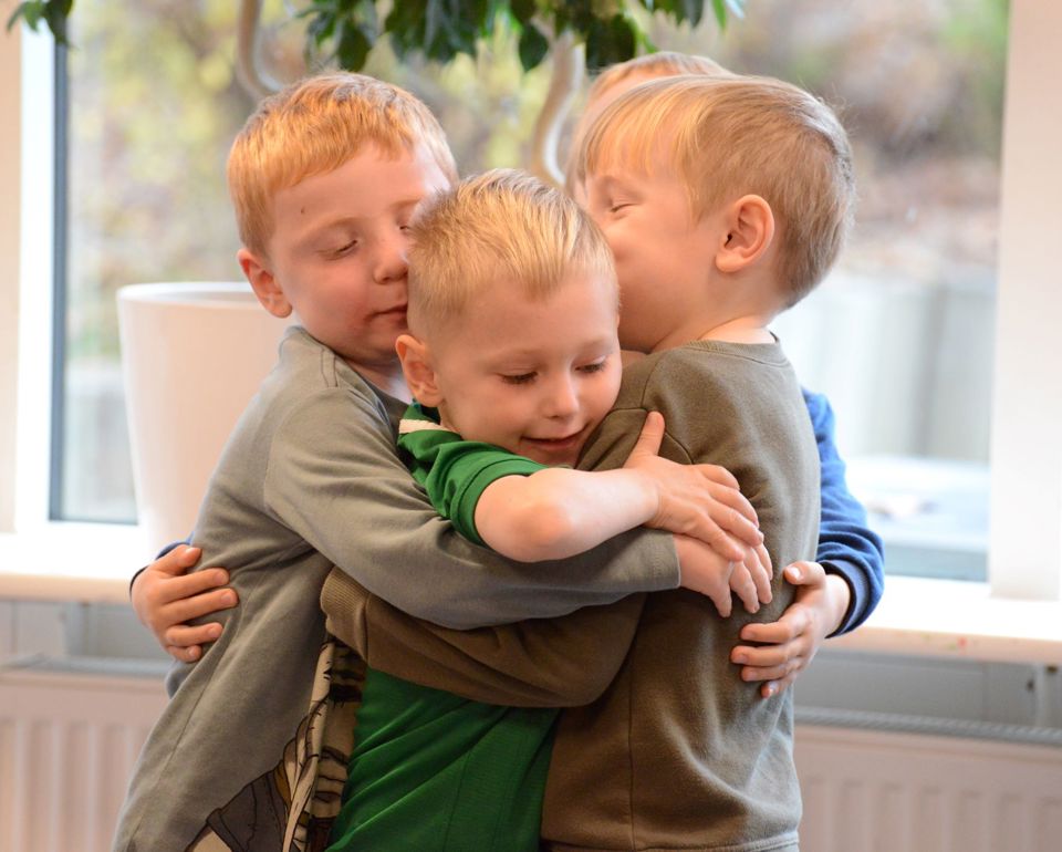 Fire børn står i et gruppekram. De er indendørs, med et vindue og noget grønt synligt i baggrunden.