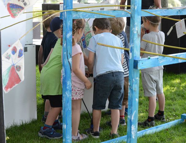 Børn samlet omkring en aktivitet inden for en blåmalet træstruktur med gule reb og dekorationer. Scenen er udendørs på græs.