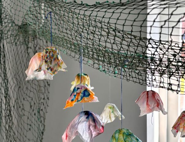 Farverige, blomsterlignende objekter hænger fra et net, der er ophængt i luften. Objekterne ser ud til at være lavet af letvægtsmateriale som papir eller stof og er farvet i forskellige farver, herunder rød, blå, grøn og gul.