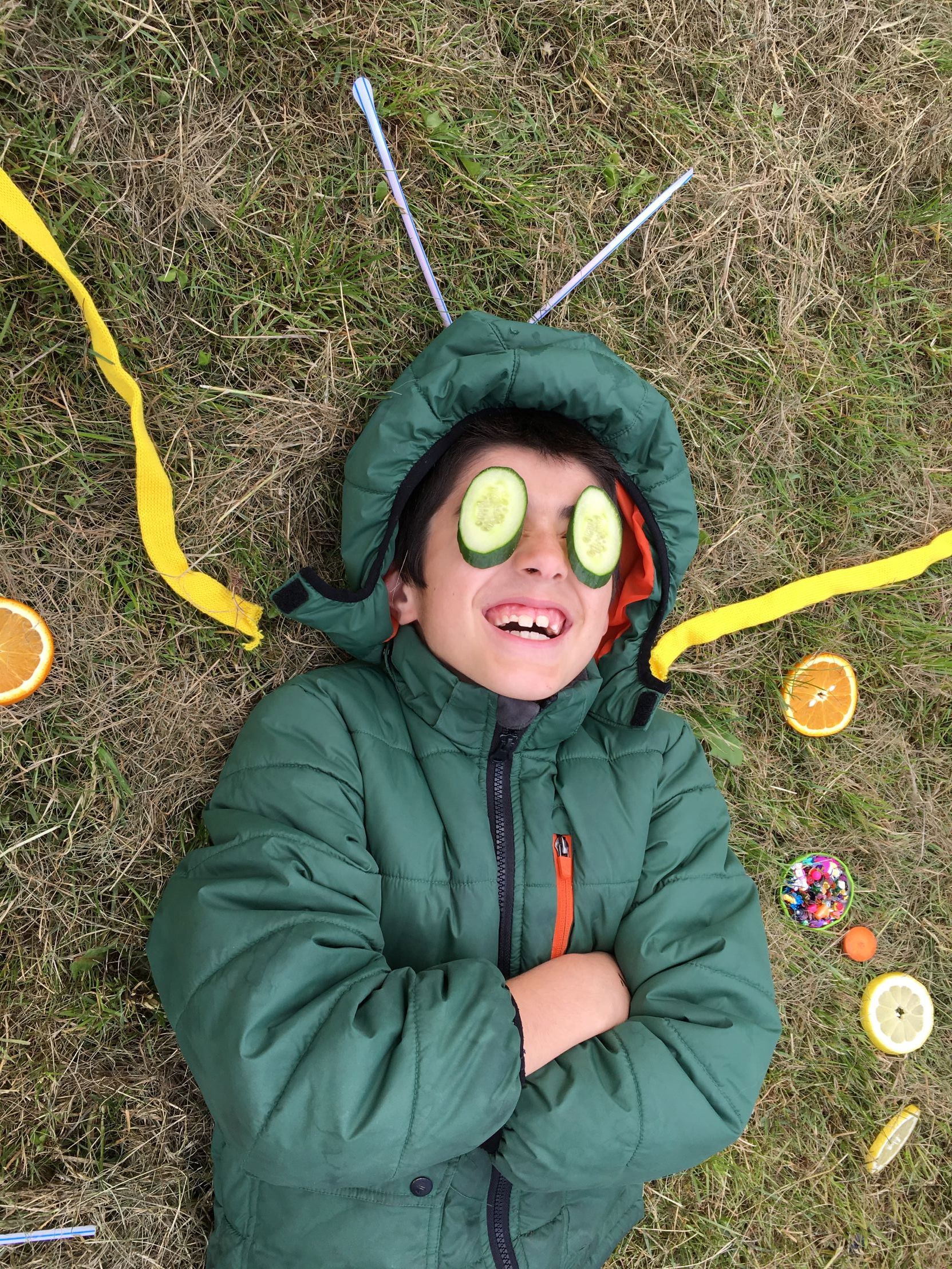 En person ligger på græsset med en grøn jakke. Der er forskellige genstande omkring personen, herunder skiver af appelsin og citron, farverige krymmel i en lille beholder, gule bånd og nogle sugerør.