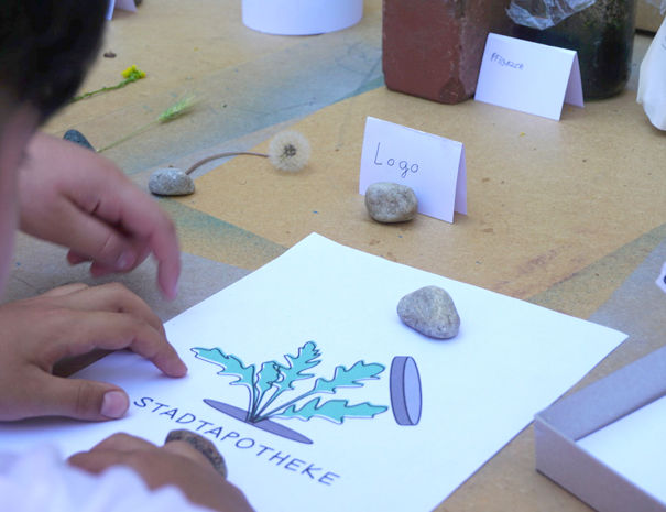 Et bord med forskellige genstande, herunder sten, papirer og etiketter. En person arbejder med et stykke papir, der har en illustration af en plante og teksten "STADTAPOTHEKE."
