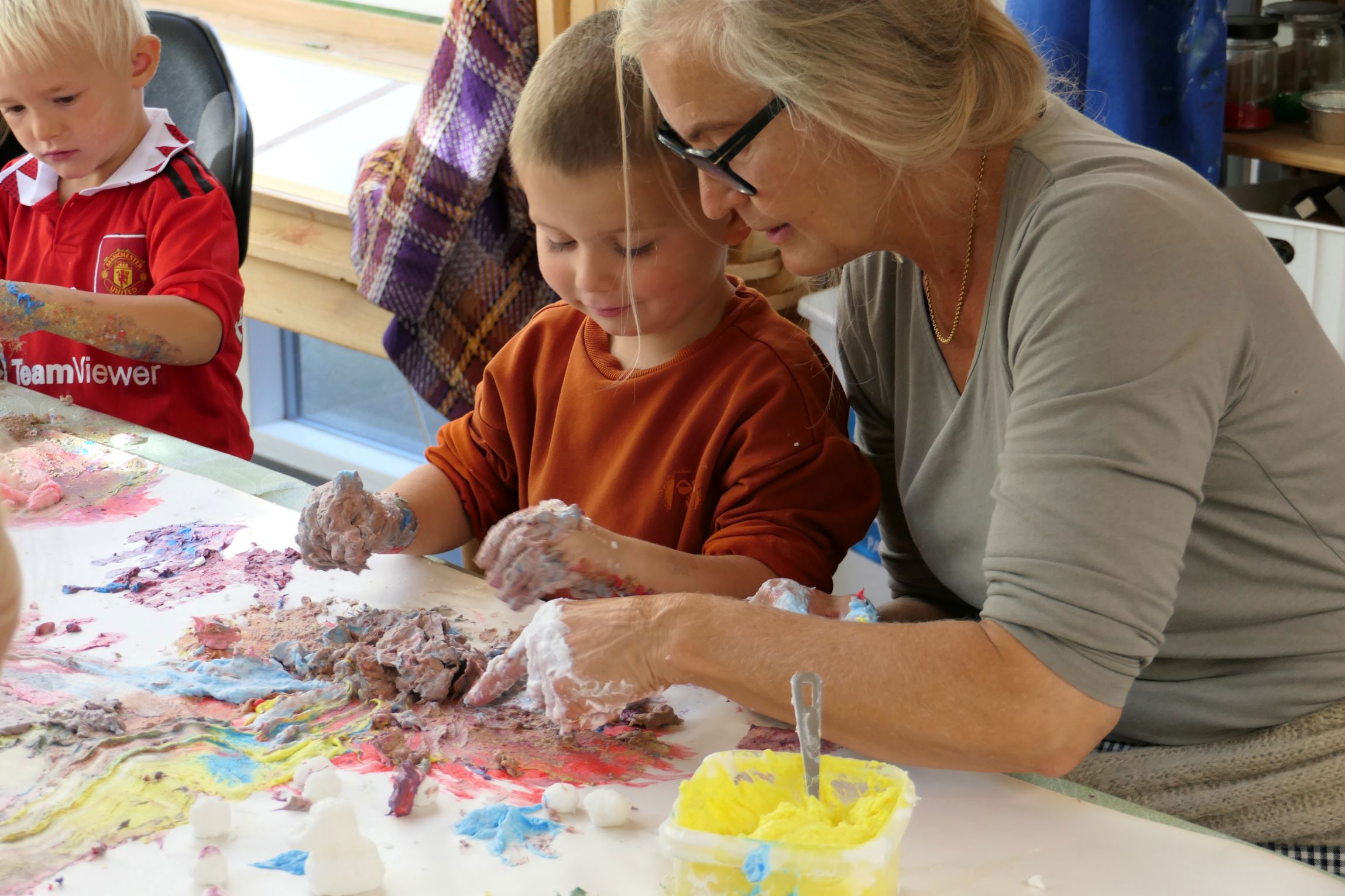 En gruppe mennesker, herunder børn og en voksen, der laver kunst ved et bord med farverige materialer. Den voksne ser ud til at hjælpe et af børnene med kunstprojektet.