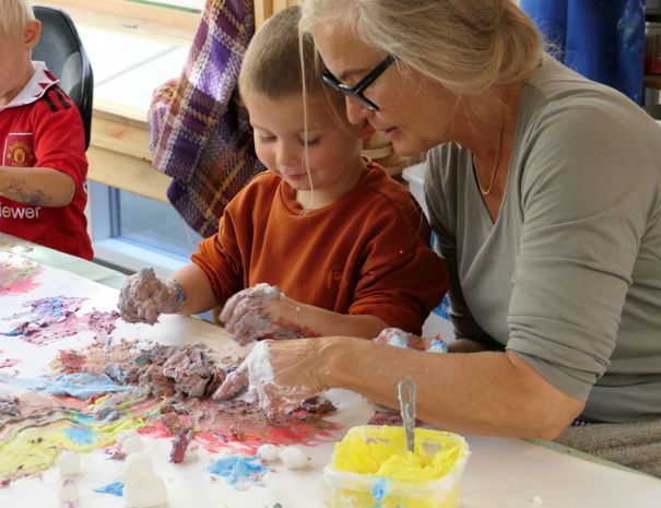 En gruppe mennesker, herunder børn og en voksen, der laver kunst ved et bord med farverige materialer. Den voksne ser ud til at hjælpe et af børnene med kunstprojektet.