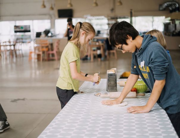 En gruppe børn er engageret i en madlavningsaktivitet. Et barn river noget med et rivejern, mens et andet barn ruller dej ud på et bord dækket med en mønstret dug.