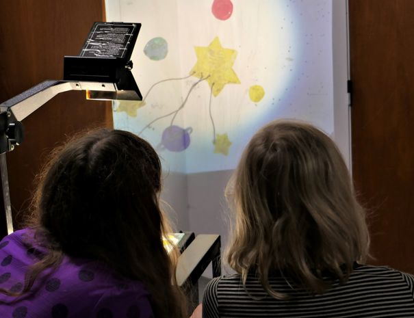 To personer sidder foran en overheadprojektor. Projektoren viser en tegning, der inkluderer flere farverige former, såsom stjerner og cirkler, på en væg.