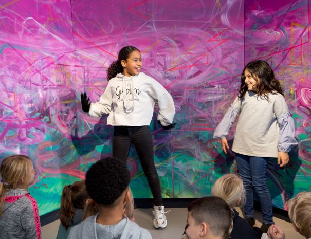 En gruppe børn står foran en farverig, abstrakt baggrund. To af børnene står op og ser ud til at være engageret i en aktivitet eller optræden, mens de andre børn sidder og kigger på.