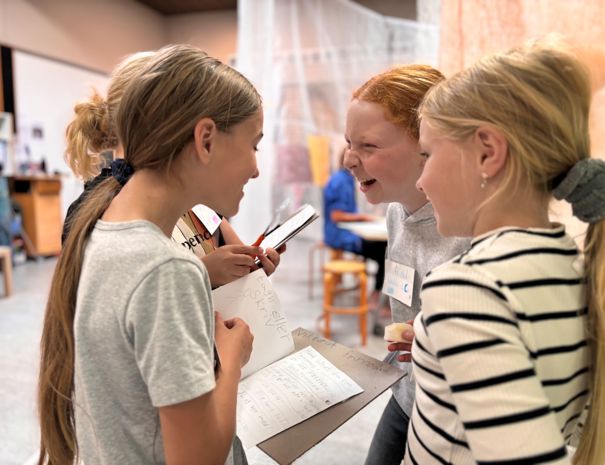 Fire personer står tæt sammen og ser ud til at være engageret i en diskussion eller samarbejdsaktivitet. De holder notesbøger og kuglepenne, hvilket tyder på, at de måske arbejder på et projekt eller lektier.