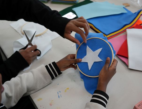 To personer laver håndarbejde. Den ene klipper hvidt stof med en saks, mens den anden holder en broderiramme med blåt stof og en hvid stjerneform på. Forskellige stykker farvet stof ligger spredt på bordet.