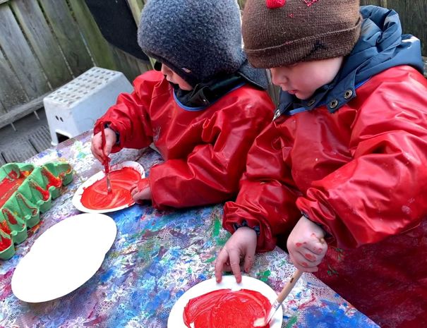 To børn iført røde forklæder og vinterhuer sidder ved et bord udendørs og maler på hvide papirtallerkener med rød maling. Bordet er dækket med en farverig, plettet dug, og der er en æggebakke malet grøn og rød på venstre side af bordet.