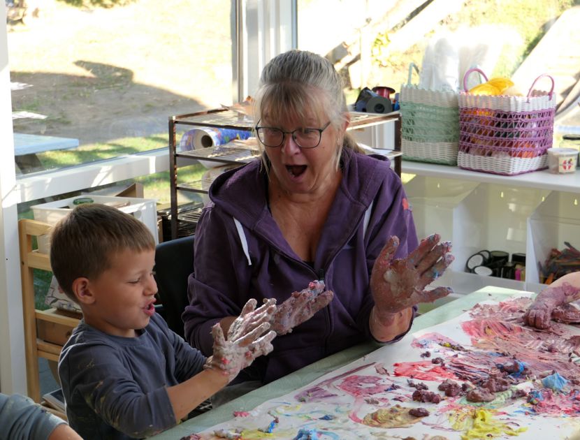 En voksen og et barn sidder ved et bord dækket med forskellige kunstmaterialer og farverig maling. Begge har malede hænder