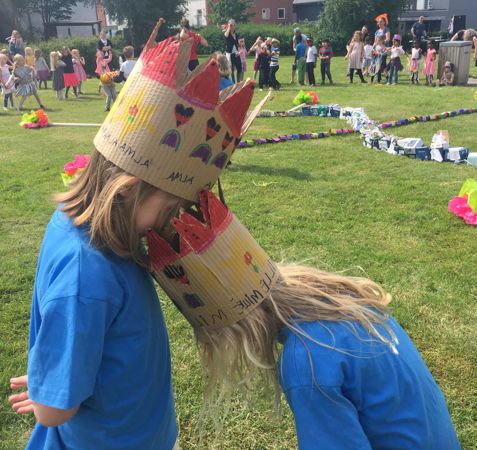 To børn med hjemmelavede kroner rører deres pander sammen i et græsareal. I baggrunden deltager andre børn og voksne i aktiviteter med forskellige dekorationer og objekter spredt rundt på marken.