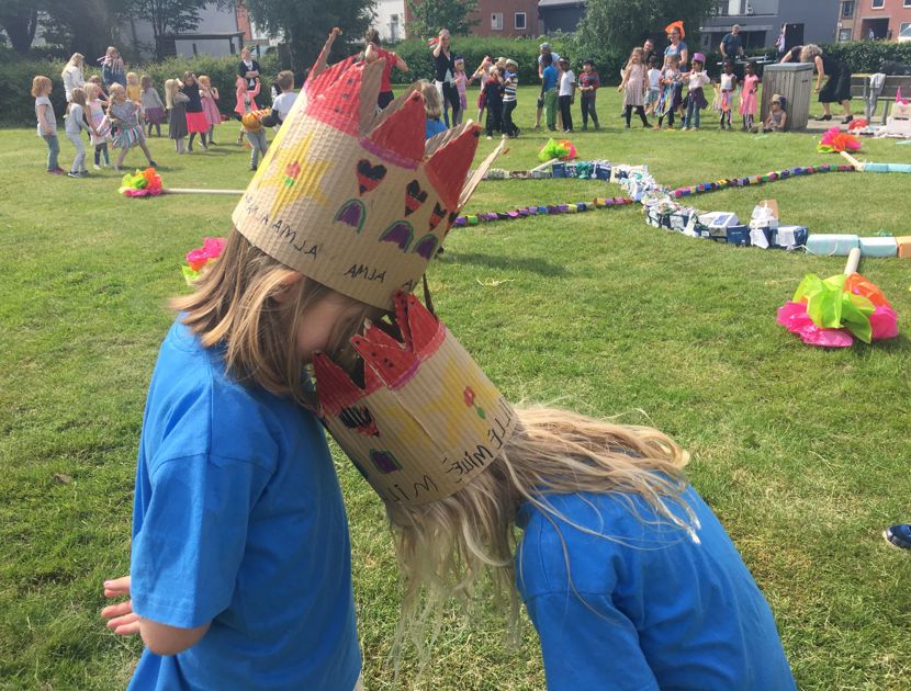 To børn med hjemmelavede kroner rører deres pander sammen i et græsareal. I baggrunden deltager andre børn og voksne i aktiviteter med forskellige dekorationer og objekter spredt rundt på marken.