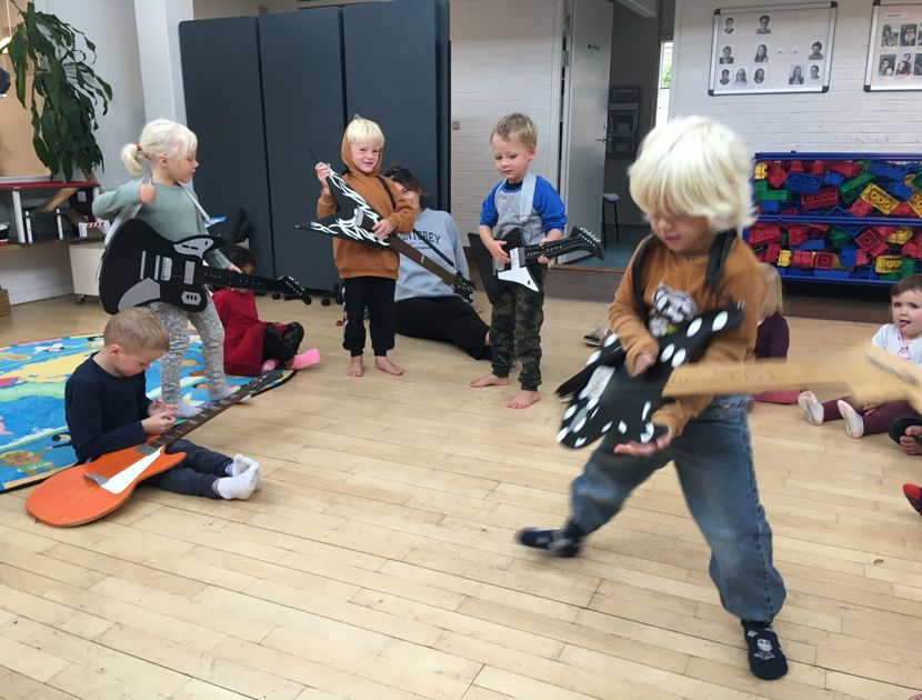 En gruppe børn leger med legetøjsguitarer i et rum. Der er fem børn synlige, hver med en legetøjsguitar, og de ser ud til at være engageret i leg. 