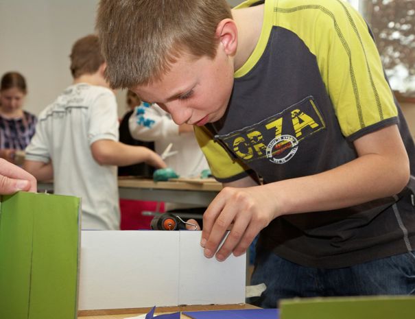 En gruppe børn engageret i en praktisk aktivitet, sandsynligvis et håndværks- eller konstruktionsprojekt. Et barn i forgrunden fokuserer på at samle stykker af pap eller lignende materiale, mens andre børn arbejder i baggrunden.