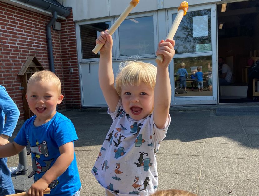 En gruppe børn leger udenfor. Et barn i midten holder to trommestikker i luften og ser ud til at være begejstret. Der er andre børn omkring, og de ser ud til at være engageret i en aktivitet sammen.