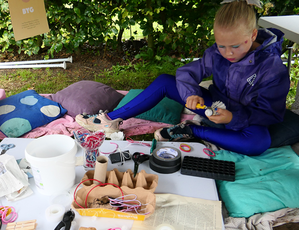 Et barn sidder på et tæppe udendørs, omgivet af forskellige håndværksmaterialer. Barnet har en lilla jakke og blå bukser på og er engageret i en form for håndværksaktivitet. Der er puder omkring barnet, og flere genstande som tape, snor, sakse, paprør og andre håndværksmaterialer er spredt ud på tæppet