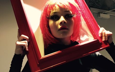 En person holder en rød billedramme foran ansigtet. Personen har rødt hår og er iført sort tøj. I baggrunden er der en dør og nogle dekorative elementer på væggen.