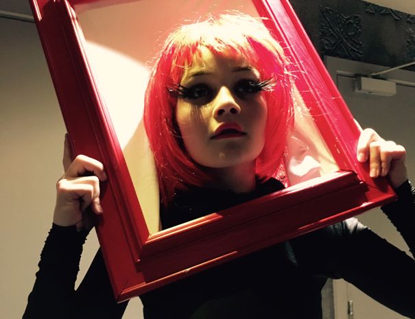 En person holder en rød billedramme foran ansigtet. Personen har rødt hår og er iført sort tøj. I baggrunden er der en dør og nogle dekorative elementer på væggen.
