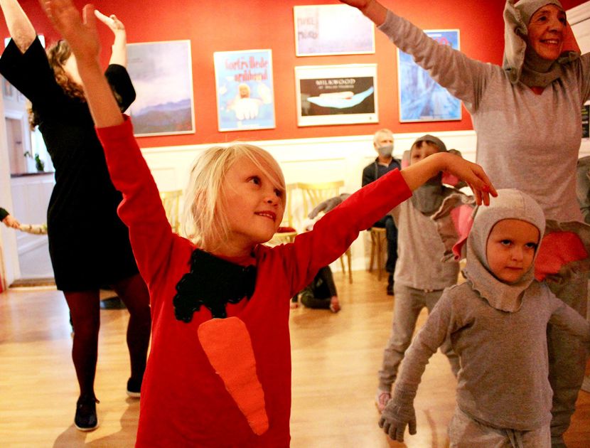En gruppe børn og en voksen i et rum med røde vægge, der deltager i en dans. Børnene er klædt i kostumer, herunder en rød trøje med en orange gulerod og grå elefant-dragter