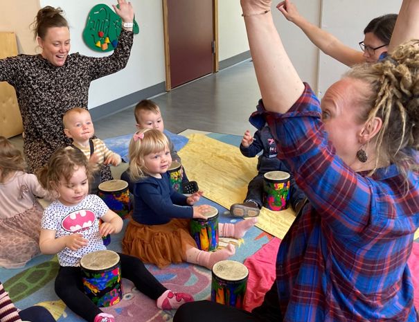 Børn og voksne sidder i en cirkel på gulvet og spiller på små trommer. En voksen i ternet skjorte har hævede arme.