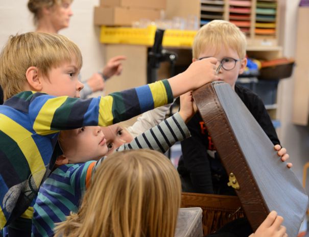 Børn samlet omkring en stor kuffert i et klasseværelse.