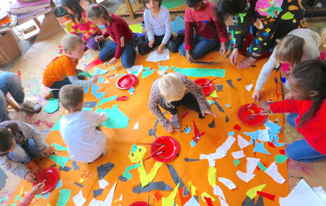 A group of children sitting on the floor around a large orange sheet of paper, creating a collage with colored paper and glue. There are red plates with glue and brushes placed around the sheet.