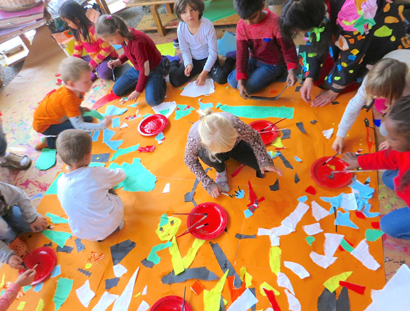 n gruppe børn sidder på gulvet omkring et stort orange ark papir og laver en collage med farvet papir og lim. Der er røde tallerkener med lim og pensler rundt om arket.