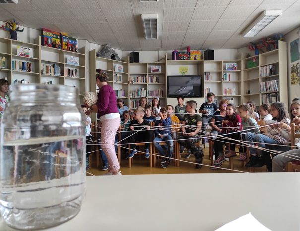 Børn sidder i en halvcirkel i et klasseværelse med en voksen stående blandt dem. Der er bogreoler fyldt med bøger i baggrunden, og børnene holder snore strakt på kryds og tværs i rummet.