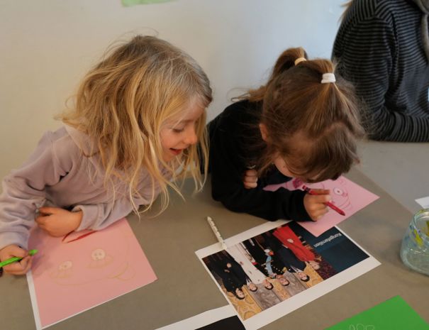 To børn sidder ved et bord og tegner. Barnet til venstre tegner på et lyserødt papir med en grøn blyant, mens barnet til højre tegner på et andet lyserødt papir med en rød blyant. Der er også et printet fotografi af kongefamilien på bordet
