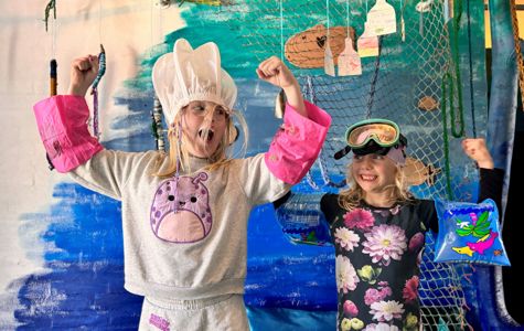 To børn står foran en blå og grøn baggrund med et fiskenet og forskellige hængende dekorationer. Begge børn har armene løftet.