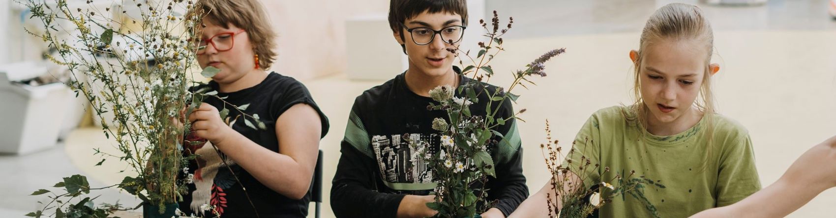 Tre børn sidder ved et bord og arbejder med planter og blomster. Bordet er dækket med forskellige plantematerialer, og der er små grønne blokke, der bruges til at arrangere planterne. En hånd rækker ind fra højre side af billedet og holder en plantestilk.