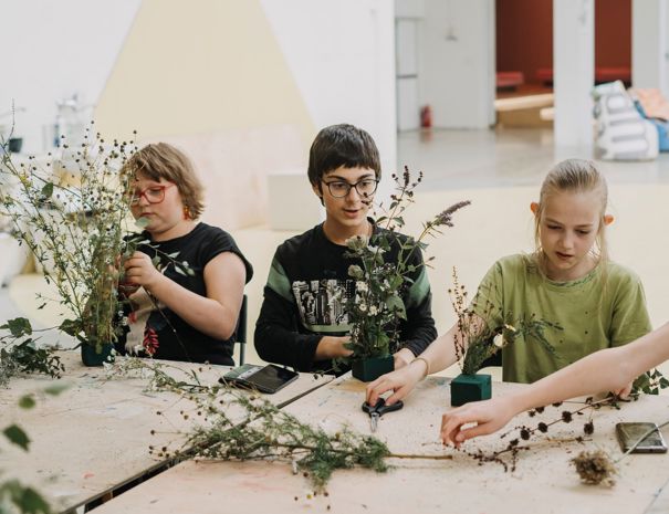 Tre børn sidder ved et bord og arbejder med planter og blomster. Bordet er dækket med forskellige plantematerialer, og der er små grønne blokke, der bruges til at arrangere planterne. En hånd rækker ind fra højre side af billedet og holder en plantestilk.