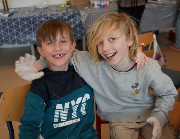 To børn sidder på stole med armene omkring hinanden. Den ene bærer en mørkeblå og grøn sweatshirt med "NYC" på, mens den anden har en grå sweatshirt med Mickey Mouse design. 