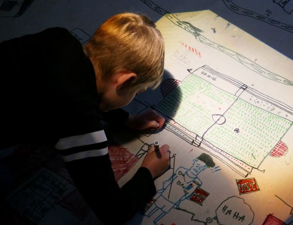 Et barn tegner på et stort stykke papir. Tegningen inkluderer forskellige elementer som en fodboldbane, mennesker og andre objekter.