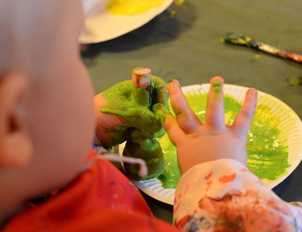 Et barn maler med fingrene og bruger grøn maling på en papirtallerken. Der er også nogle pensler i baggrunden.