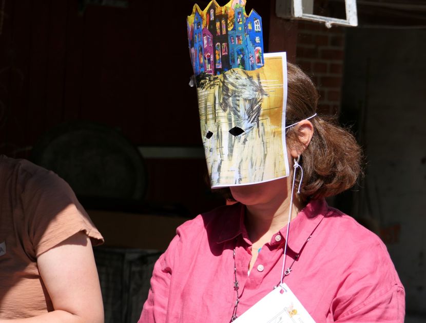 En person med en maske, der dækker hele ansigtet og er dekoreret med farverige tegninger af bygninger. Personen har en lyserød skjorte på og en navneskilt om halsen.