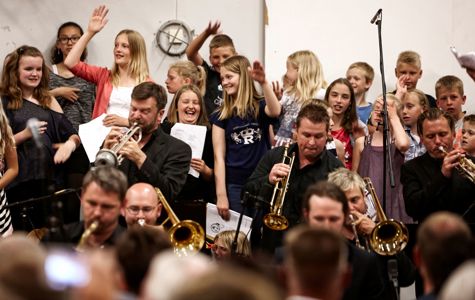 En gruppe mennesker, herunder børn og voksne, i en musikalsk optræden eller øvelse. Børnene står og holder nogle papirark, mens de voksne sidder og spiller messinginstrumenter som trompeter og tromboner.