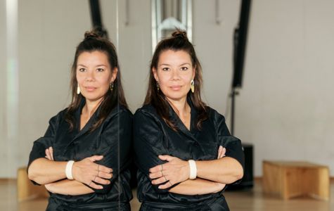 Camilla Høg stands with arms crossed in front of a mirror, with her reflection visible. The background includes some wooden furniture and structural elements of the room.