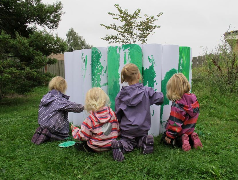 En gruppe børn sidder på græsset og maler på store hvide cylindriske objekter med grøn maling. Scenen ser ud til at være udendørs, med træer og et hegn i baggrunden.