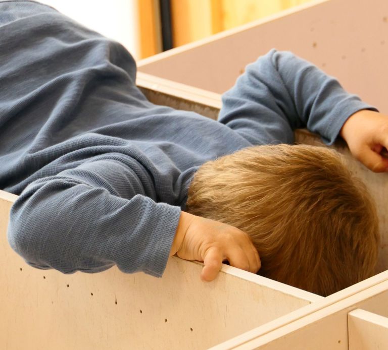 t barn med lysebrunt hår iført en blå langærmet trøje læner sig over og kigger ind i en tom kasse, med begge hænder på kanten.